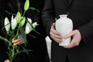 two people standing in black clothes at funeral, one is holding white lilies and the other is holding a white urn