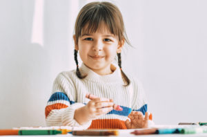 Happy cute little girl paints with oil pencils, sitting at white desk at home. Pretty smiling preschool kid draws with colorful pencils.