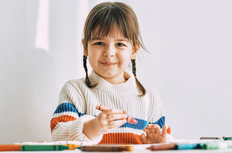 Happy cute little girl paints with oil pencils, sitting at white desk at home. Pretty smiling preschool kid draws with colorful pencils.