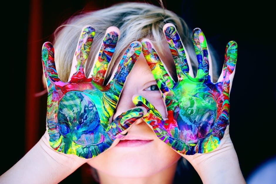 blond haired child, holding hands covered in paint in front of face with only one eye visible