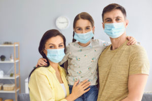 Family in medical masks on the face looks at the camera while standing in the room at home.